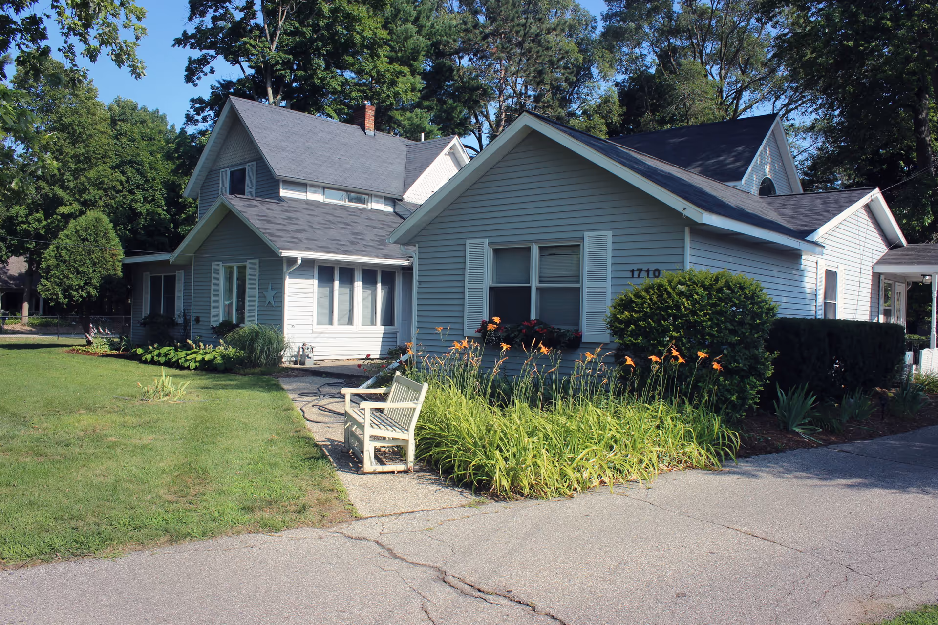 Front exterior of a gray siding residential-style building with lawn, flower beds, and a bench by the driveway.