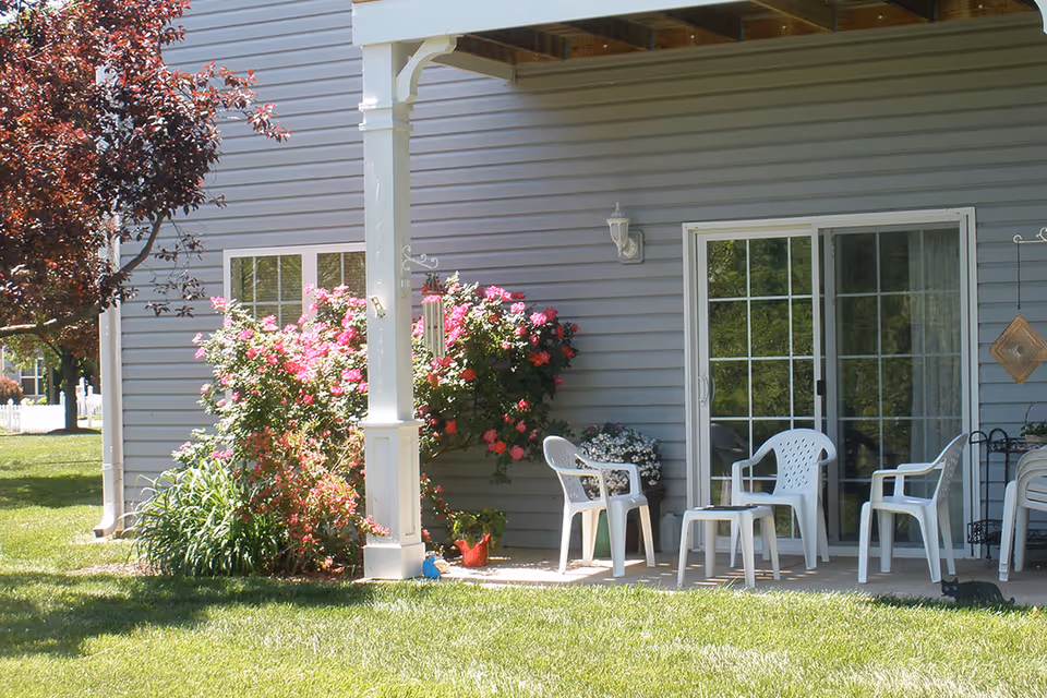 A covered patio area outside a building with gray siding, featuring white plastic chairs and a small table. There are pink flowering bushes and other plants near the patio, with a green lawn in the foreground and a tree with reddish leaves on the left side.