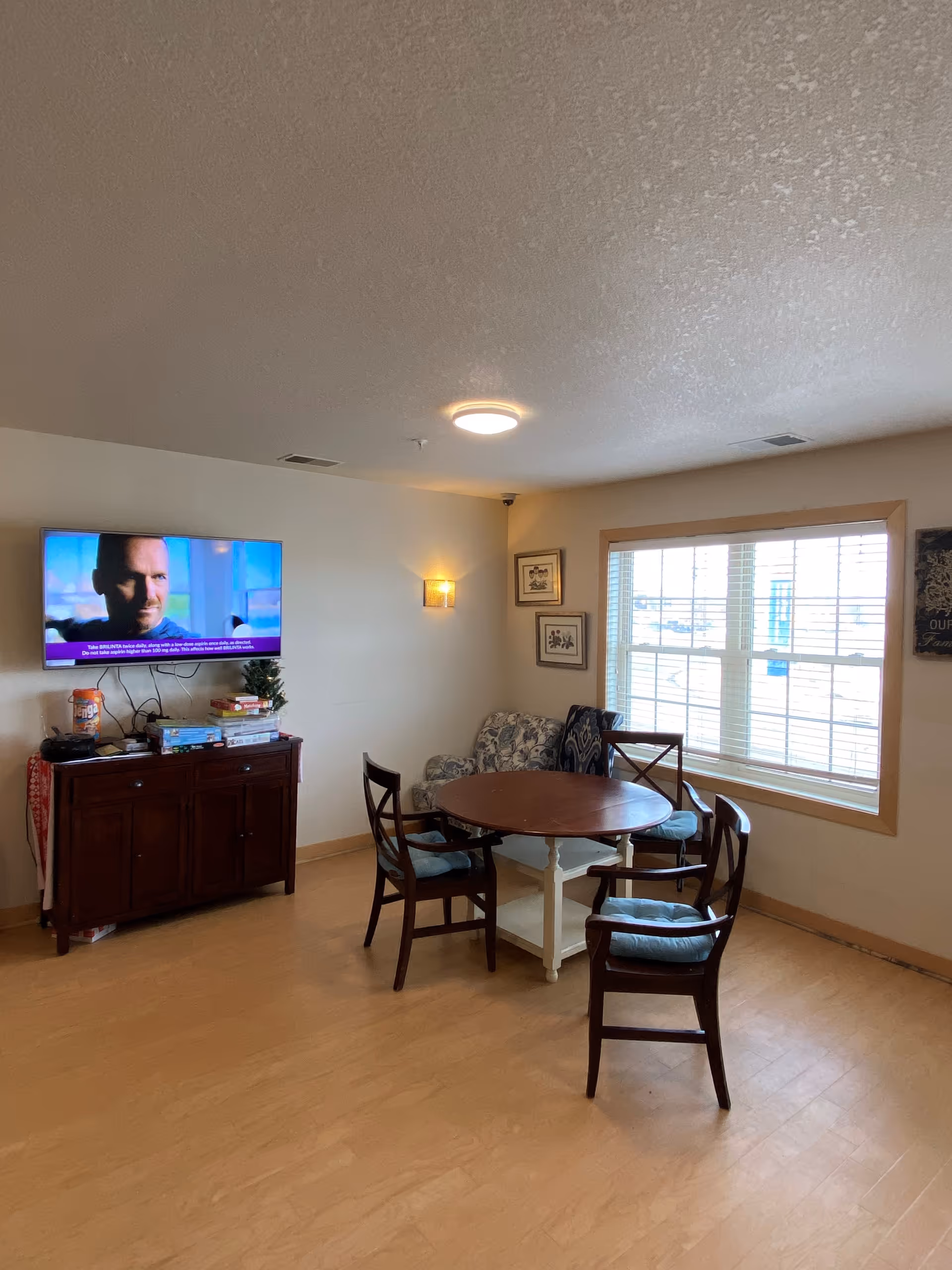 Bright senior living common room with a round table and four chairs, a TV on a cabinet, an upholstered chair and a large window.