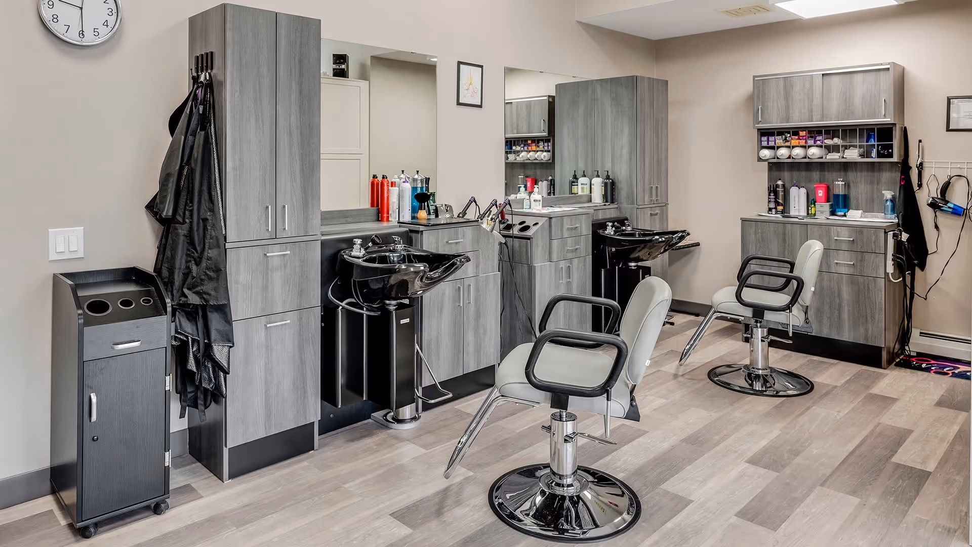 Interior view of a hair salon area in a senior living facility with two styling chairs, two black hair washing sinks, grey cabinets, various hair care products on the counters, and a wall clock.