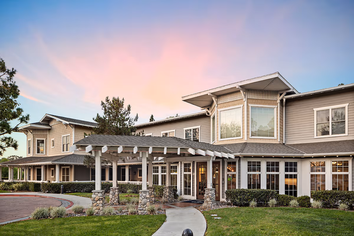 Exterior view of a two-story senior living facility building with a covered entrance supported by stone pillars, surrounded by landscaped greenery and a curved walkway, under a colorful sunset sky.