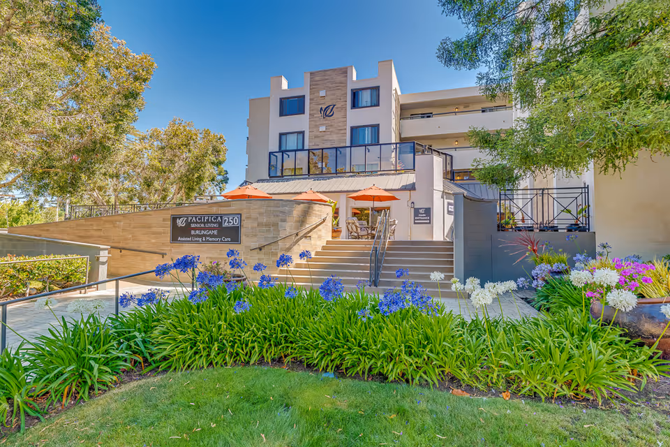 Front entrance of Burlingame Senior Living building with steps, outdoor seating umbrellas and flowering landscaping.