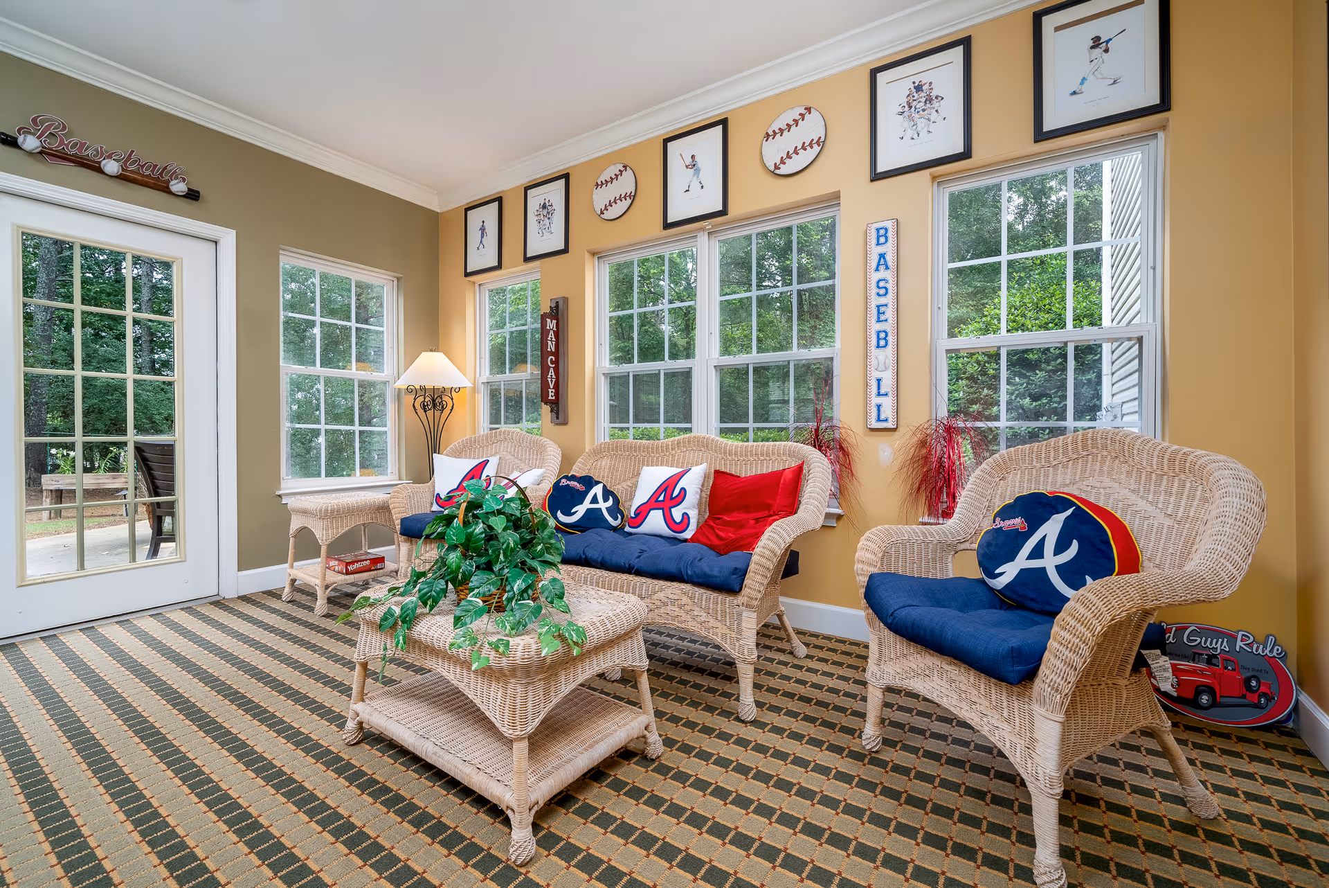 A cozy sunroom with wicker furniture including a loveseat, two chairs, and a coffee table. The furniture has blue cushions and pillows with the Atlanta Braves logo. The walls are decorated with baseball-themed artwork and signs, including framed pictures of baseball players and baseballs. Large windows let in natural light and show greenery outside. A floor lamp stands in the corner, and a patterned carpet covers the floor.