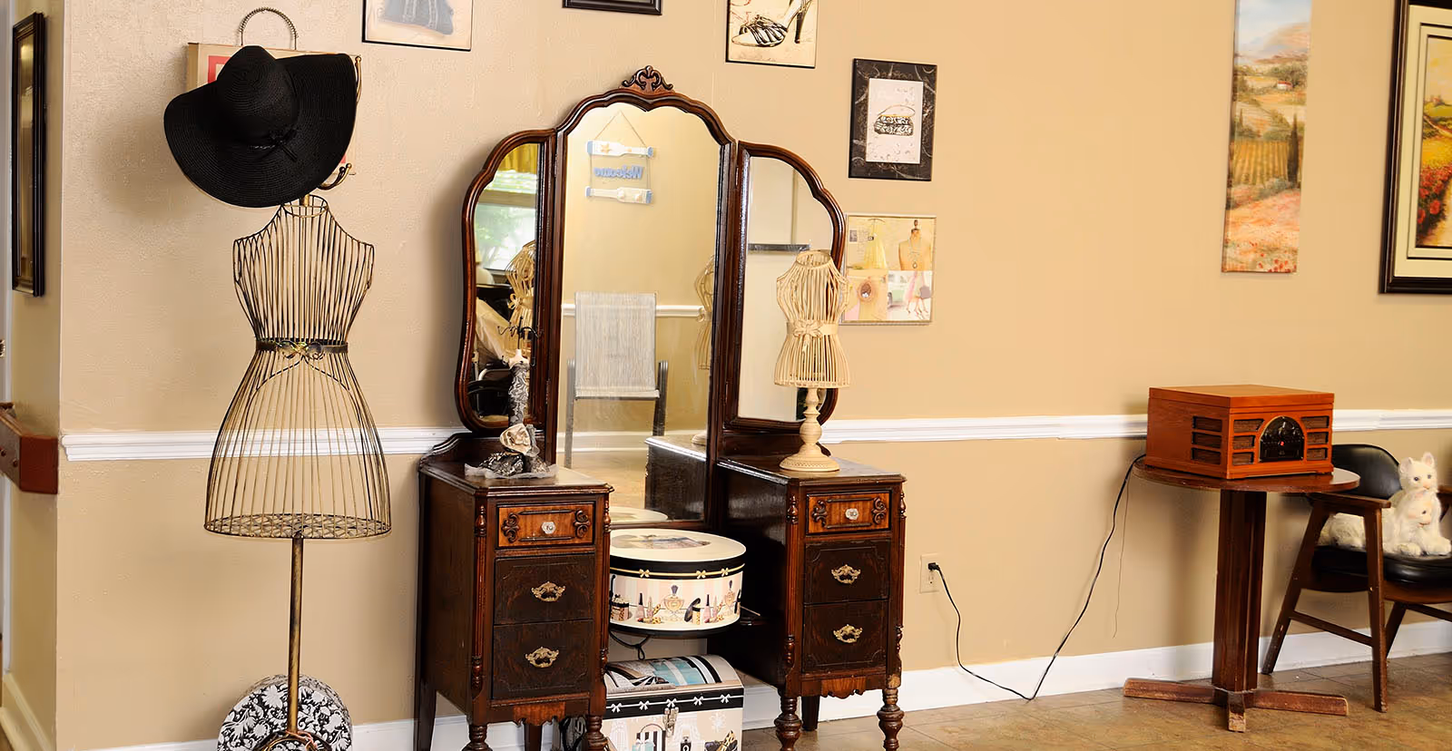 A cozy interior featuring a vintage dressing table with a tri-fold mirror, a wire dress form wearing a black hat, and a side table with a record player.