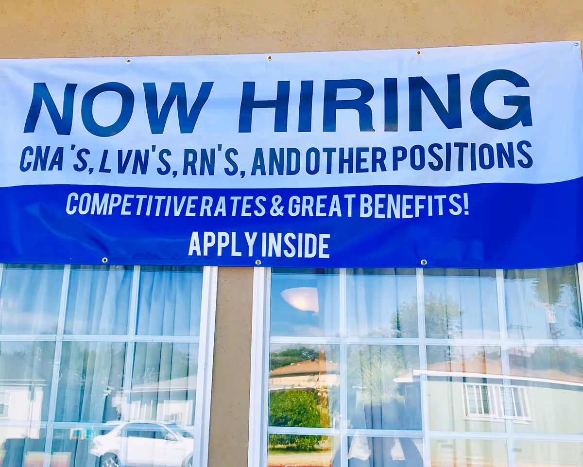 A large banner hanging on the exterior wall of a building with the text 'NOW HIRING CNA'S, LVN'S, RN'S, AND OTHER POSITIONS COMPETITIVE RATES & GREAT BENEFITS! APPLY INSIDE'. Below the banner, there are windows reflecting cars and houses outside.