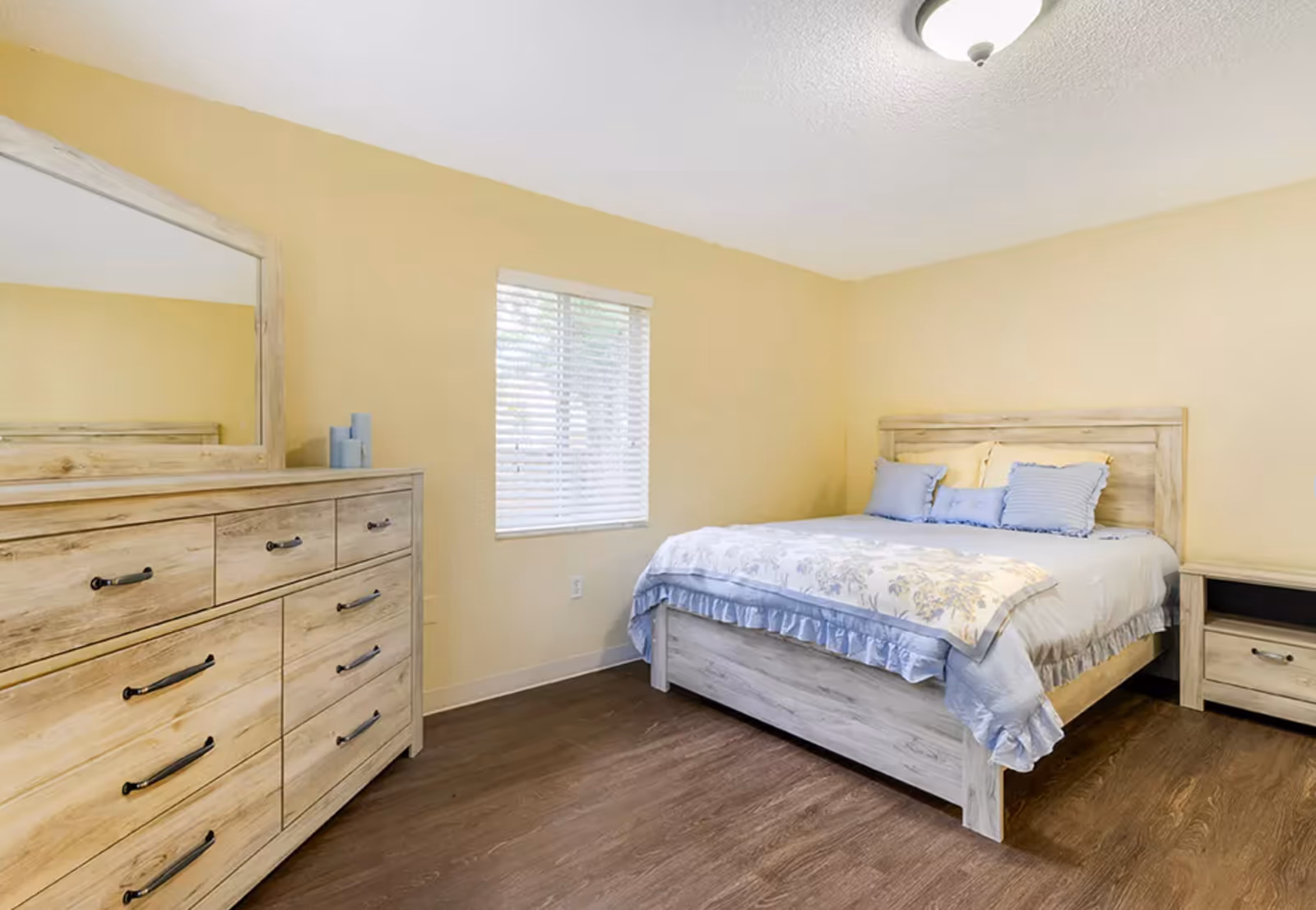 A bedroom with light wood furniture including a bed with blue and yellow pillows and a floral bedspread, a dresser with a large mirror, and a nightstand. The walls are painted light yellow and there is a window with white blinds letting in natural light. The floor is a dark wood laminate.