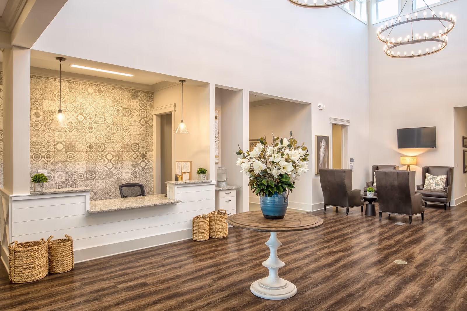 A spacious and well-lit senior living facility lobby with a reception desk on the left featuring a patterned tile wall and two hanging pendant lights. In the center, there is a round wooden table with a large blue vase filled with white flowers. To the right, there is a seating area with four dark upholstered chairs, a small table, a floor lamp, and a wall-mounted TV. The floor is wood with a rich brown finish, and the walls are painted white with high ceilings and large circular chandeliers.