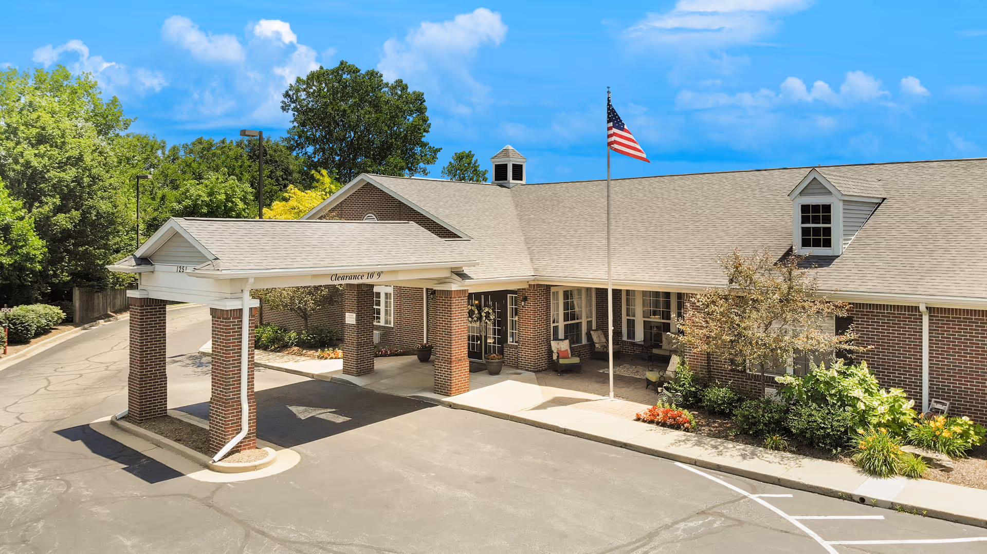 Exterior view of a single-story brick building with a covered entrance supported by brick columns. An American flag is flying on a flagpole near the entrance. The building is surrounded by greenery and trees under a blue sky with some clouds.