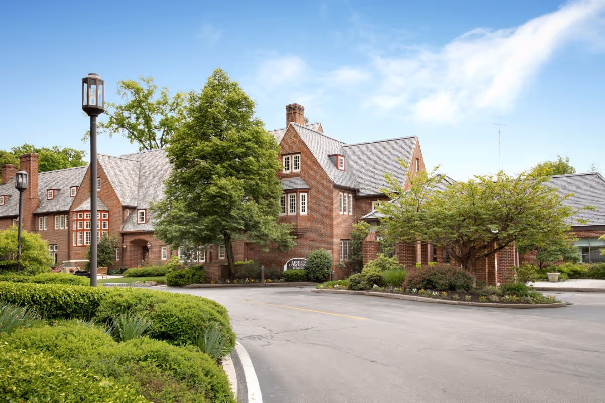 Exterior view of a large brick building with multiple windows and a steep gray roof, surrounded by green trees and bushes under a partly cloudy blue sky. A curved driveway leads up to the entrance, and there are street lamps along the path.
