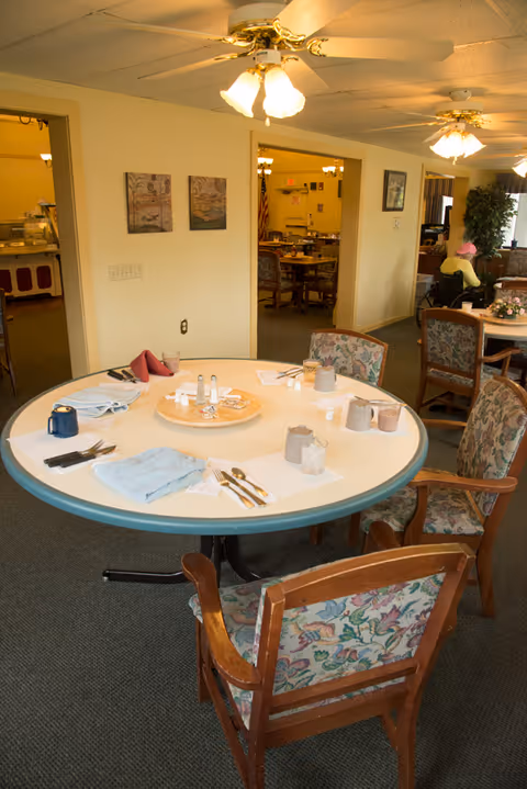 Round dining table set with utensils, napkins, salt and pepper shakers, and condiments in a senior living facility dining room with floral upholstered chairs. Ceiling fans with lights hang overhead, and additional tables and chairs are visible in adjoining rooms.