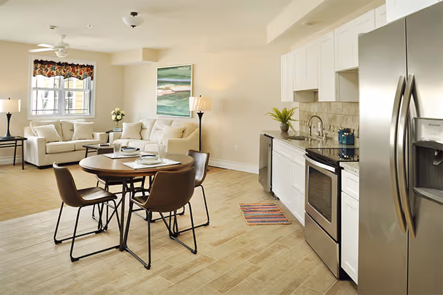 A bright and modern open-concept living space featuring a kitchen with stainless steel appliances, white cabinets, and granite countertops on the right. In the center, there is a round dining table set with four brown chairs and place settings. On the left side, a cozy living area with two beige sofas, a coffee table, a floor lamp, and a window with a floral valance is visible. The walls are light-colored, and a large abstract painting hangs above one sofa.