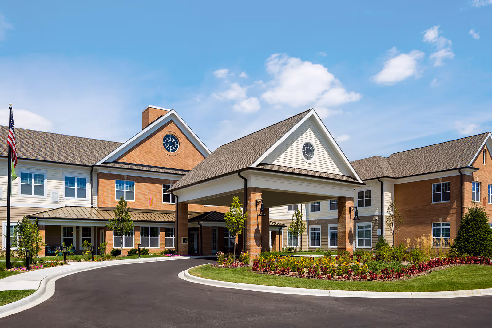 Exterior view of Arbor Terrace Glenview, a senior living facility with a large covered entrance, brick and siding facade, well-maintained landscaping, and a clear blue sky with some clouds.