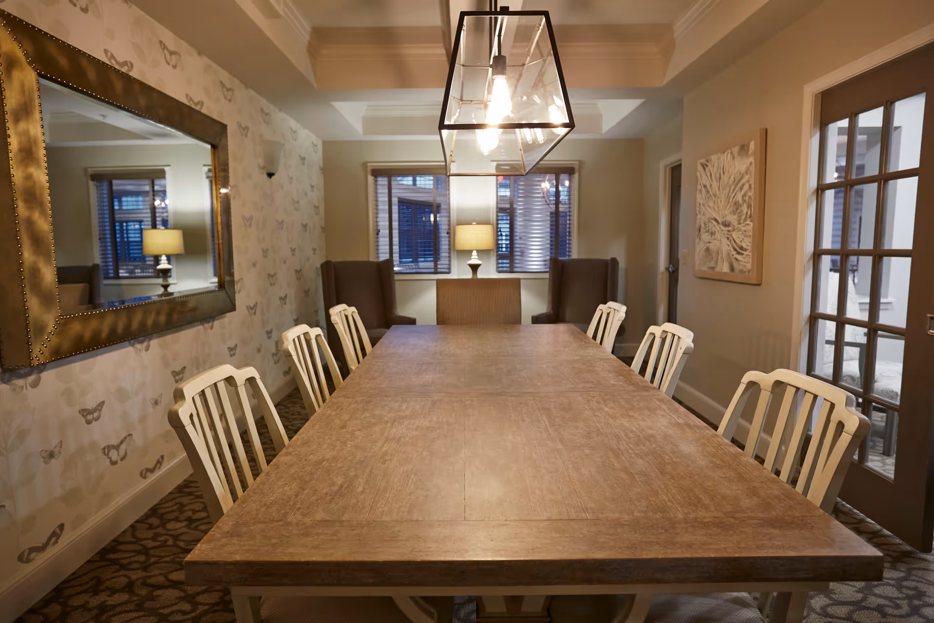 Long wooden dining table with white chairs under a rectangular pendant light in a warmly lit dining room with a large mirror and windows.