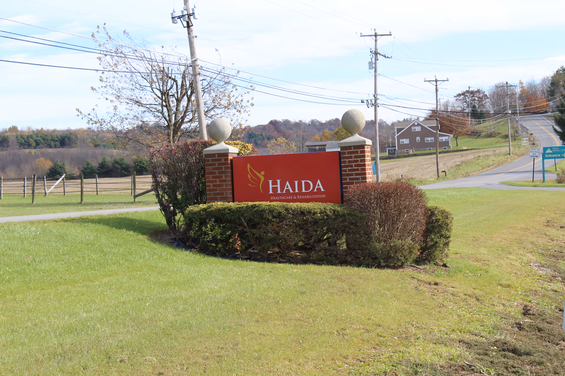 Outdoor view of a red sign with white text reading 'HAIDA Healthcare & Rehabilitation' set between two brick pillars with spherical stone tops, surrounded by trimmed bushes and grass. In the background, there is a rural road, utility poles, a house, and trees with autumn foliage under a partly cloudy sky.