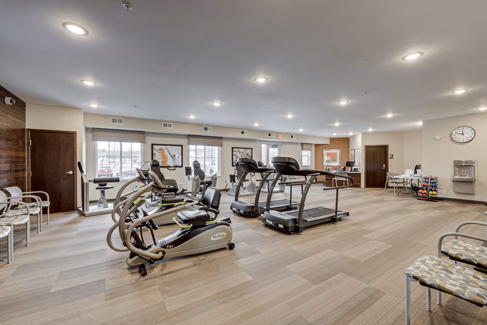 A spacious fitness room in a senior living facility with exercise equipment including treadmills, recumbent bikes, and weight racks. The room has large windows letting in natural light, beige walls, and patterned carpet flooring. Several chairs are lined up along the walls, and a clock and water fountain are visible on the right wall.