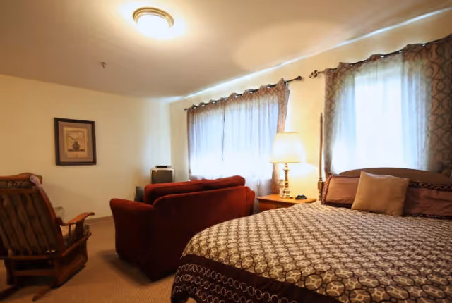 A cozy bedroom with a large bed covered in a patterned bedspread, a wooden rocking chair, a red sofa, a wooden nightstand with a lamp, and two windows with curtains letting in soft light.