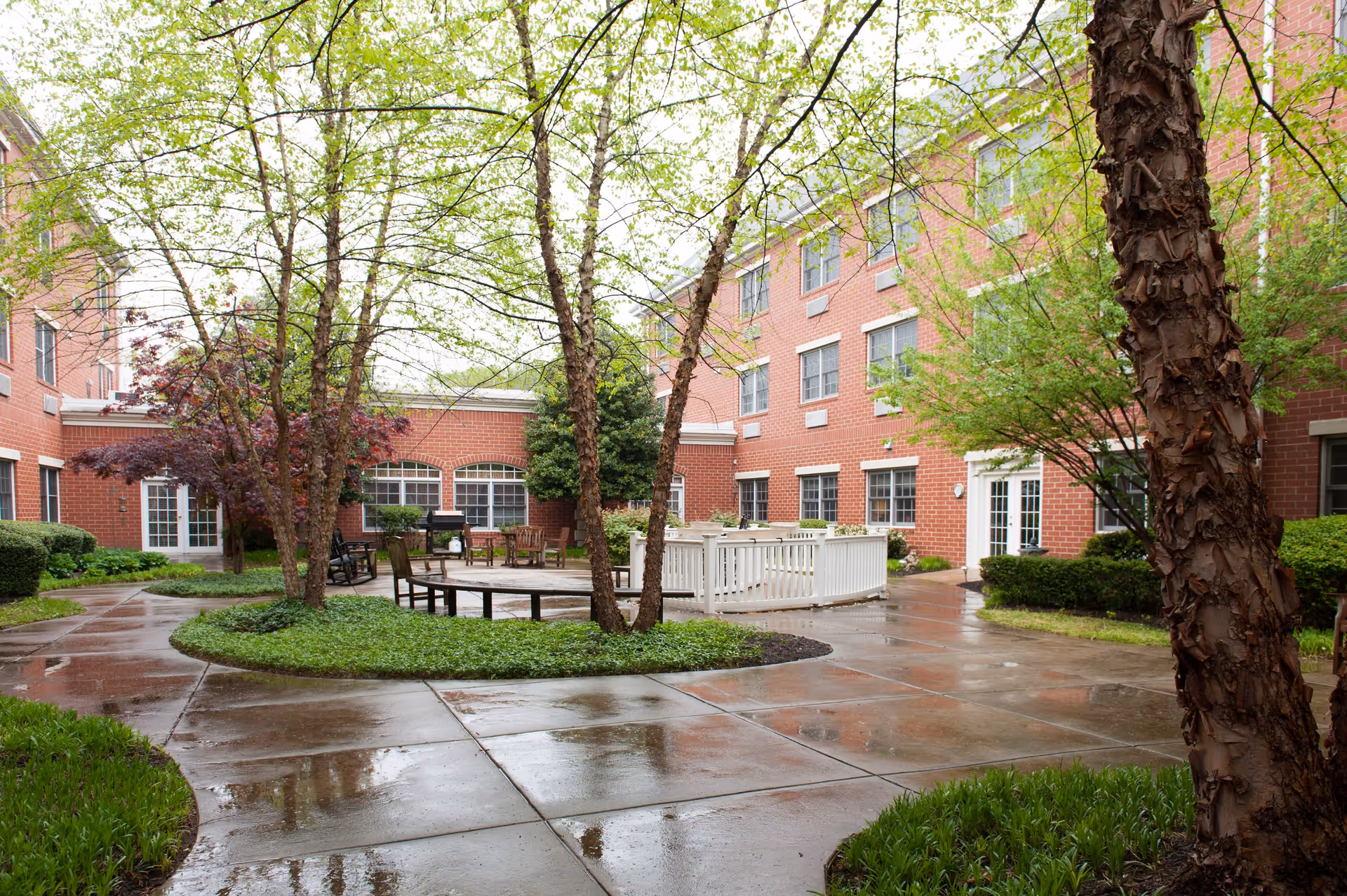 Outdoor courtyard area of a senior living facility with wet concrete walkways, green trees, shrubs, and seating areas including benches and chairs. The surrounding building is made of red brick with multiple windows and white doors.