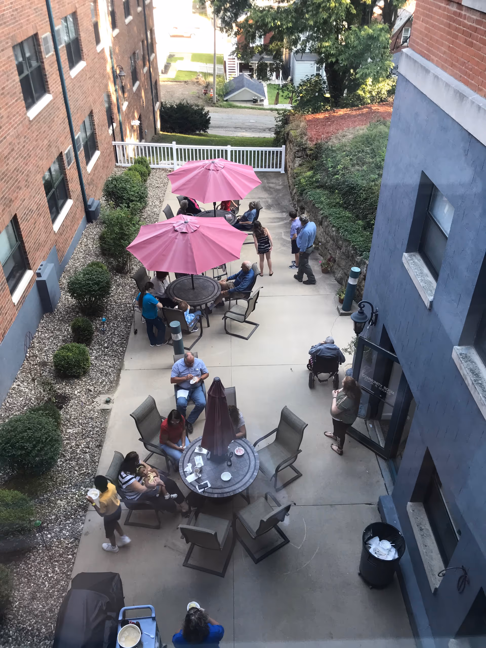 Outdoor patio area at Ahva Living of East Dubuque with several people sitting and standing around tables with pink umbrellas. The patio is surrounded by brick and blue buildings, with some greenery and a white fence in the background.