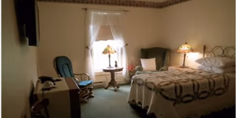 A cozy bedroom with a single bed covered in a patterned quilt, two bedside tables each with a decorative lamp, a green upholstered armchair, a small round table in front of a window with sheer white curtains, and a television mounted on the wall.