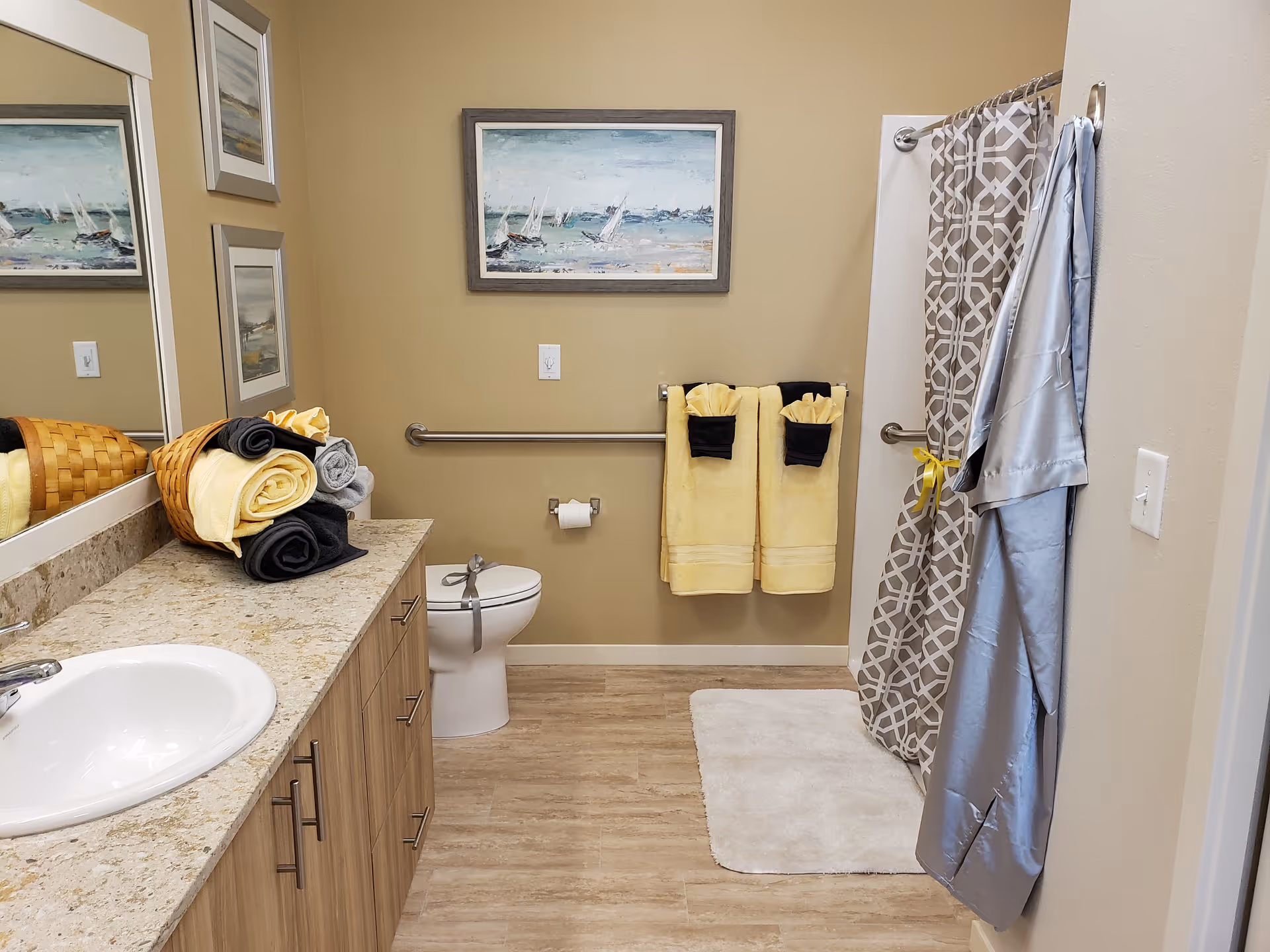 A clean and well-organized bathroom with a sink and countertop on the left, a toilet in the center, and a shower with a patterned curtain on the right. There are neatly folded yellow, black, and gray towels on the countertop and hanging on the wall. A bathrobe hangs on a hook near the shower. The walls are beige, and there are framed paintings above the toilet and on the adjacent wall.