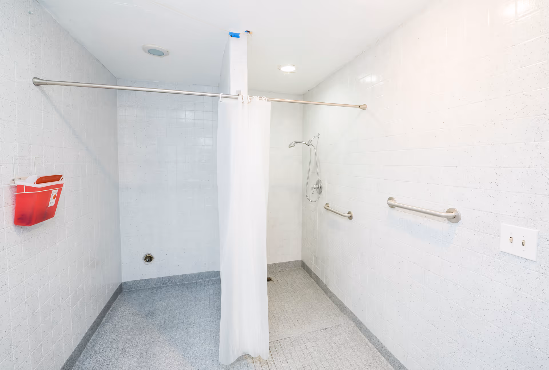 A clean, accessible shower area with white tiled walls and gray tiled floor. The shower has a white curtain hanging from a curved rod, a handheld showerhead, and two grab bars on the wall for support. There is a red wall-mounted container on the left side and a light switch on the right wall.