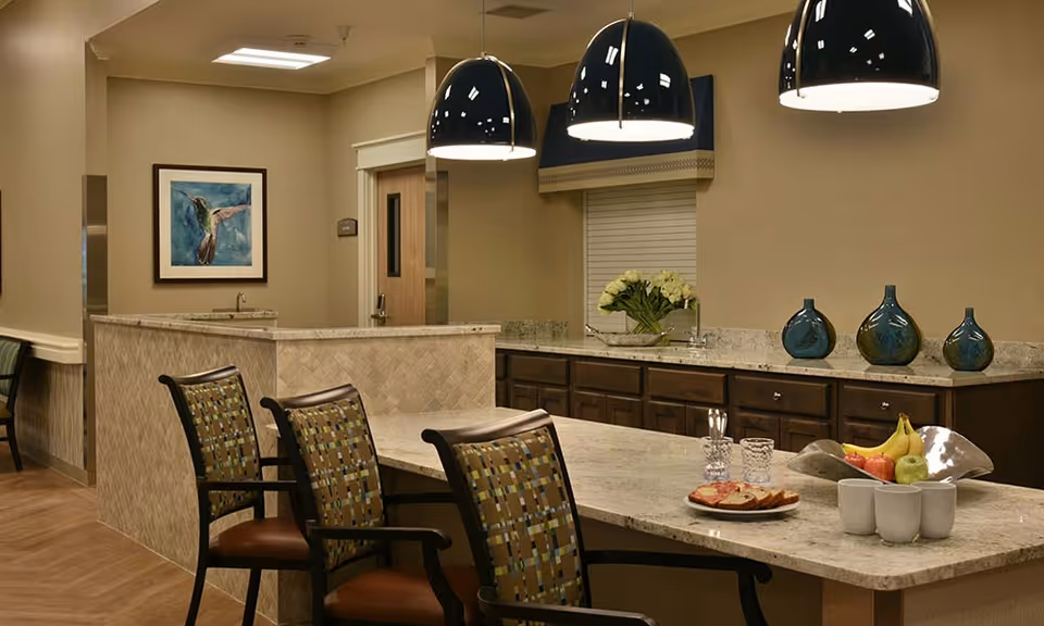 A well-lit communal dining and kitchen area with a marble countertop, chairs, pendant lamps, decorative vases and a bowl of fruit.