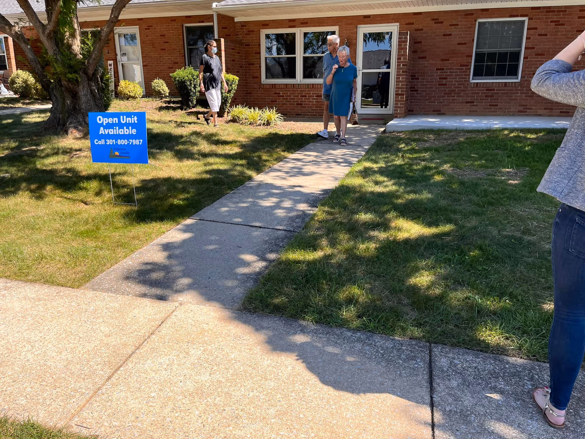 An elderly couple walking on a sidewalk away from a brick building with a woman standing nearby wearing a mask. There is a blue sign on the grass that reads 'Open Unit Available Call 301-800-7987'. Another person is partially visible on the right side of the image.