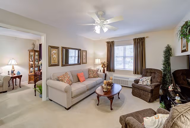 A cozy living room with a beige sofa adorned with patterned and solid pillows, a wooden coffee table with decorative items, two brown armchairs, a ceiling fan with lights, framed artwork on the walls, and a window with brown curtains letting in natural light. Adjacent room visible with additional seating and a display cabinet.
