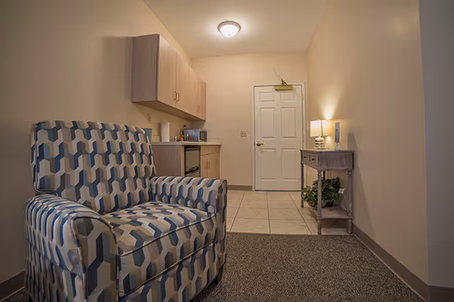 Small living area with a patterned armchair, kitchenette cabinets, entry door, and a console table with a lamp.