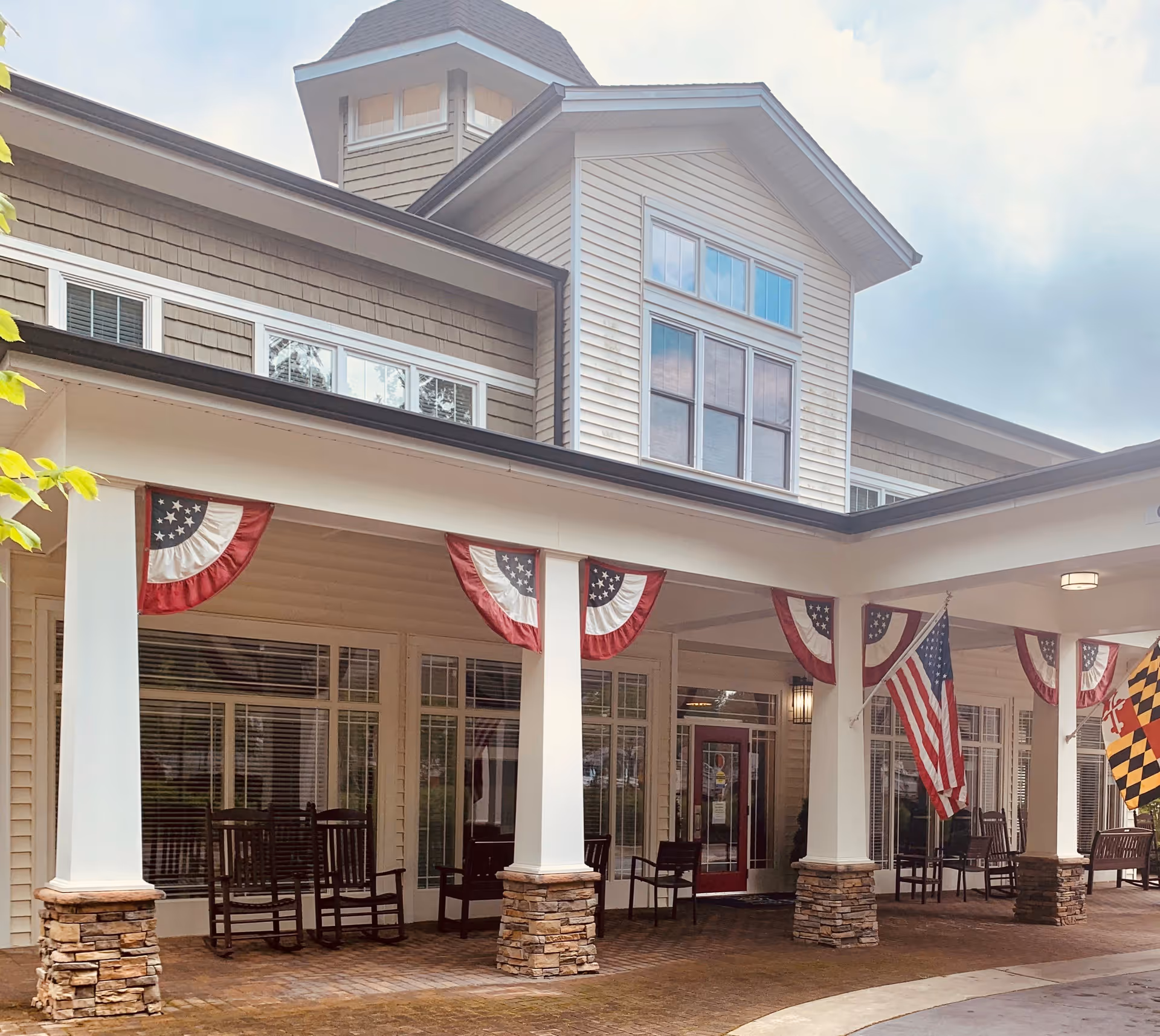 Front exterior view of a senior living facility with a covered entrance supported by white columns with stone bases. The building has beige siding and multiple windows. American patriotic bunting and flags are displayed along the entrance area. Several wooden rocking chairs are placed under the covered porch.