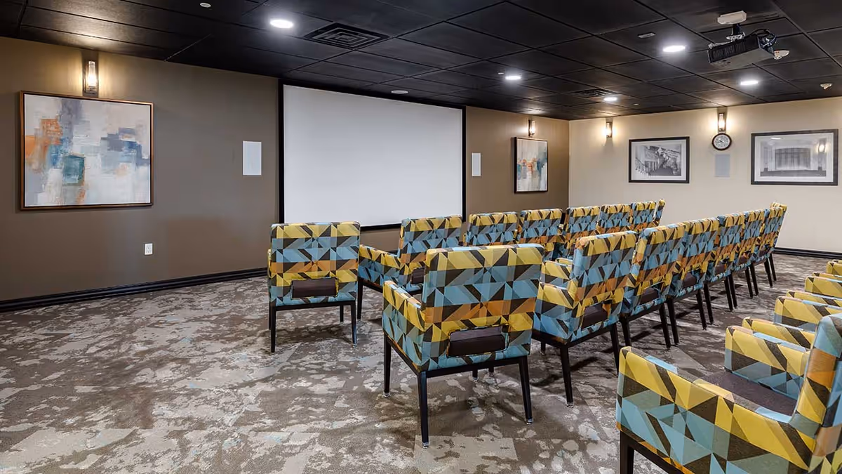 A small theater room with rows of colorful patterned chairs facing a large white projection screen. The room has a carpeted floor, brown walls with framed artwork, and a clock on the wall. Ceiling lights illuminate the space.