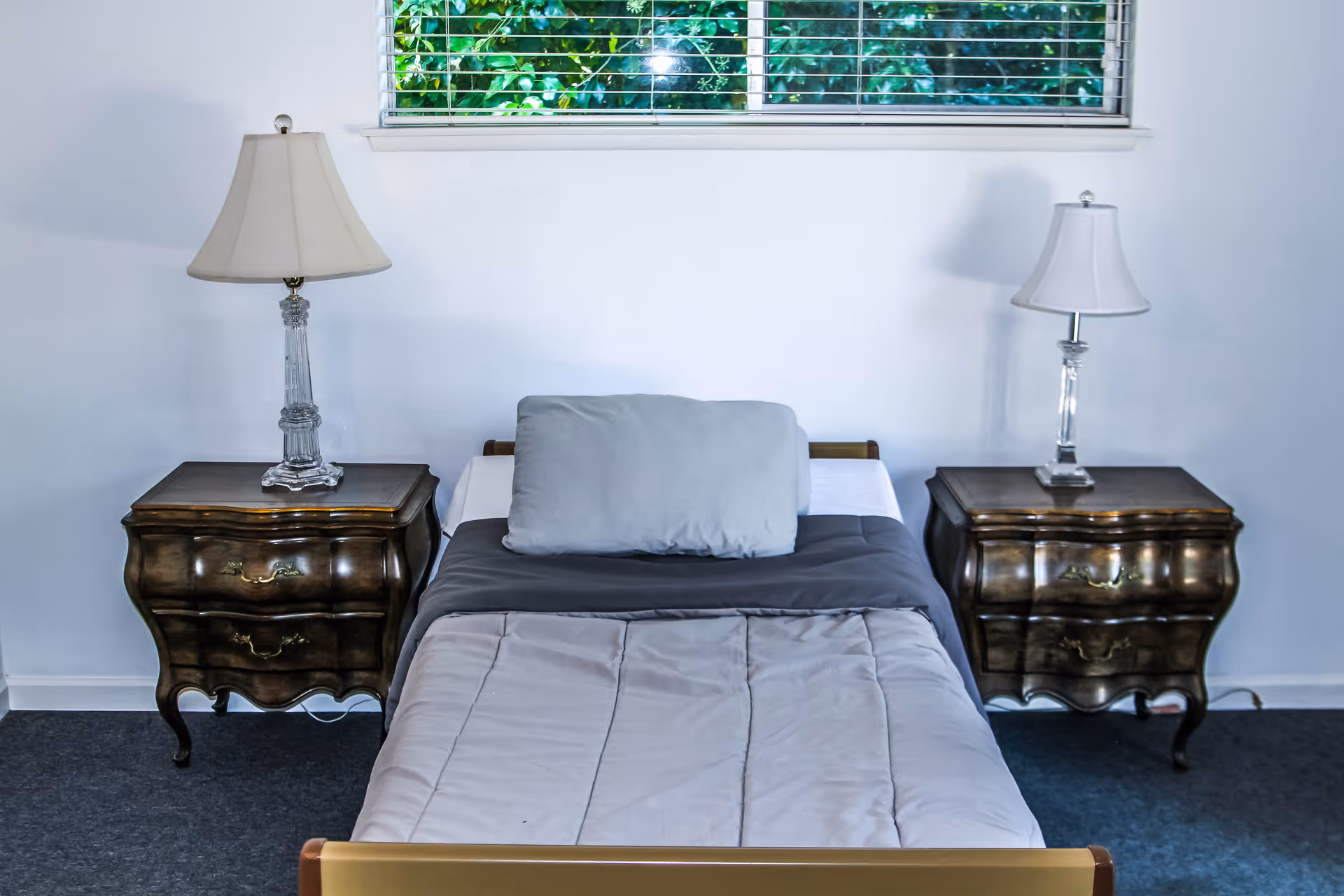A single bed with a gray pillow and light gray comforter is centered between two vintage wooden nightstands, each topped with a clear glass lamp with a white lampshade. A window with white blinds and green foliage outside is above the bed.