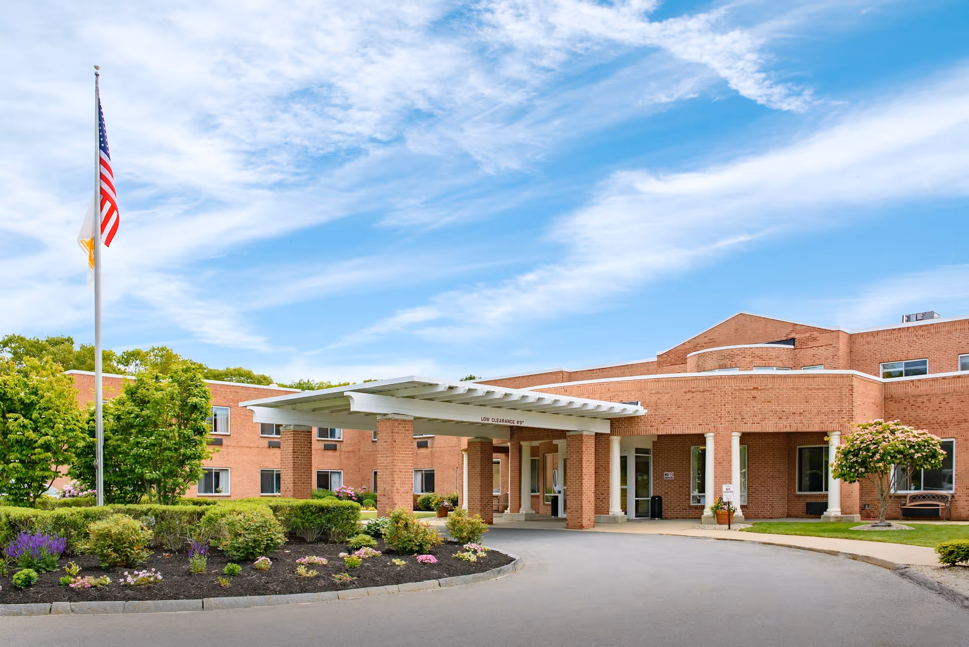 Front entrance of a brick senior living facility with a covered porte-cochère, landscaped beds, and an American flag.