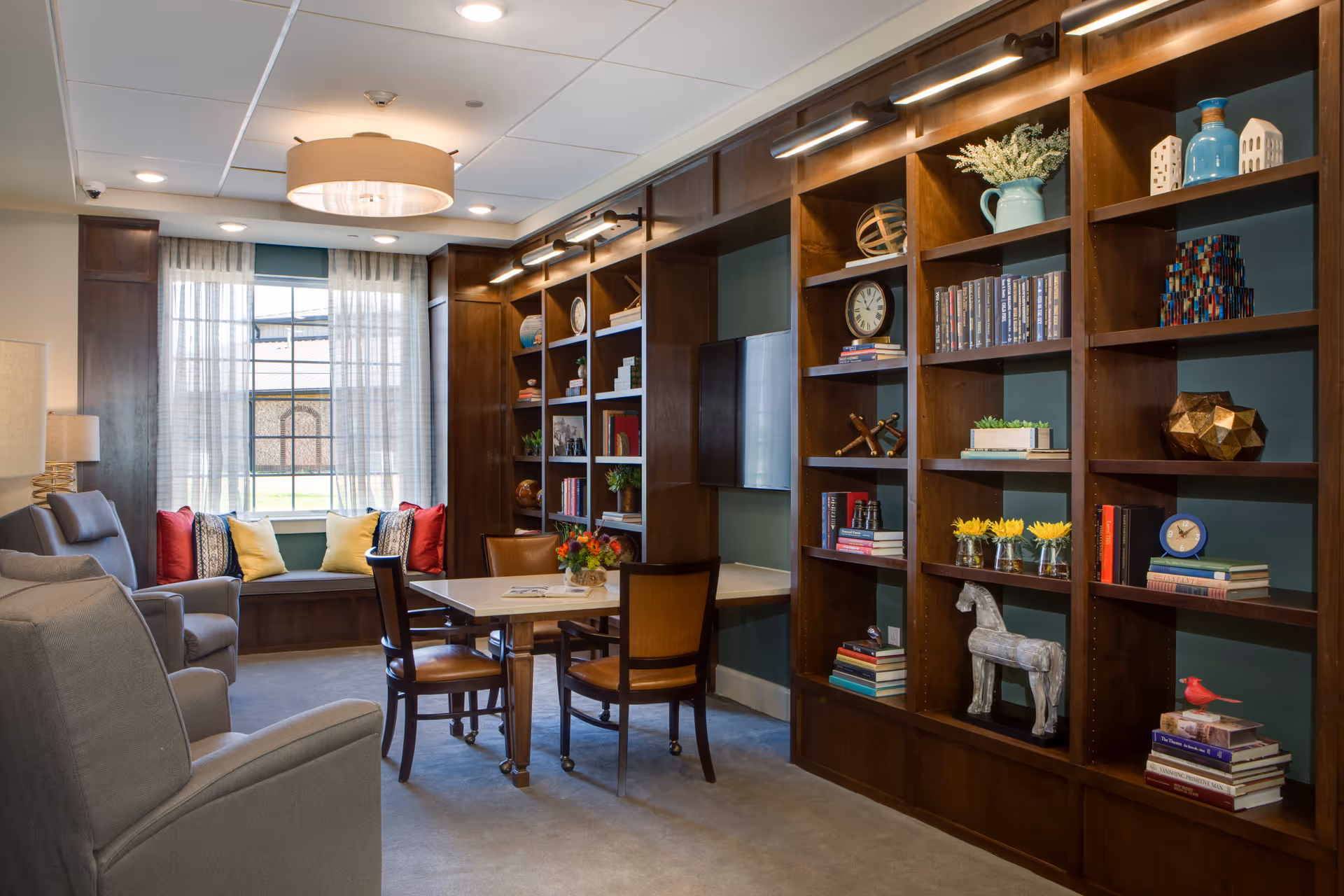 A cozy living room area with a large wooden bookshelf filled with books, decorative items, and a mounted TV. There is a table with four chairs in the center, two gray armchairs on the left, and a window seat with colorful cushions under a window with sheer curtains. The room is softly lit with ceiling lights and wall-mounted lights above the bookshelf.