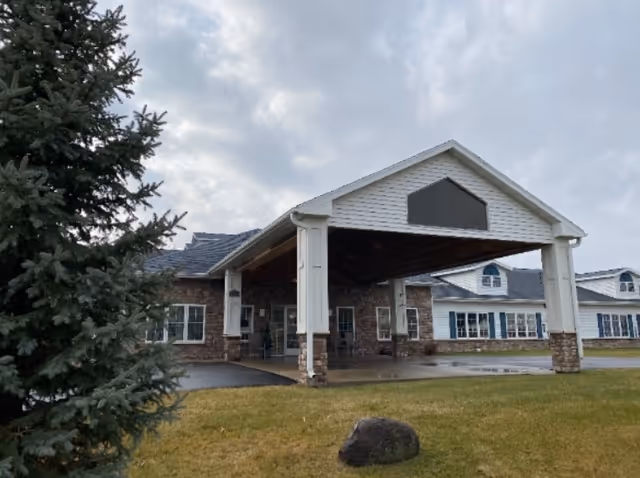 Front entrance of a single-story assisted living building featuring a covered porte-cochere, grass lawn and an evergreen tree under a cloudy sky.