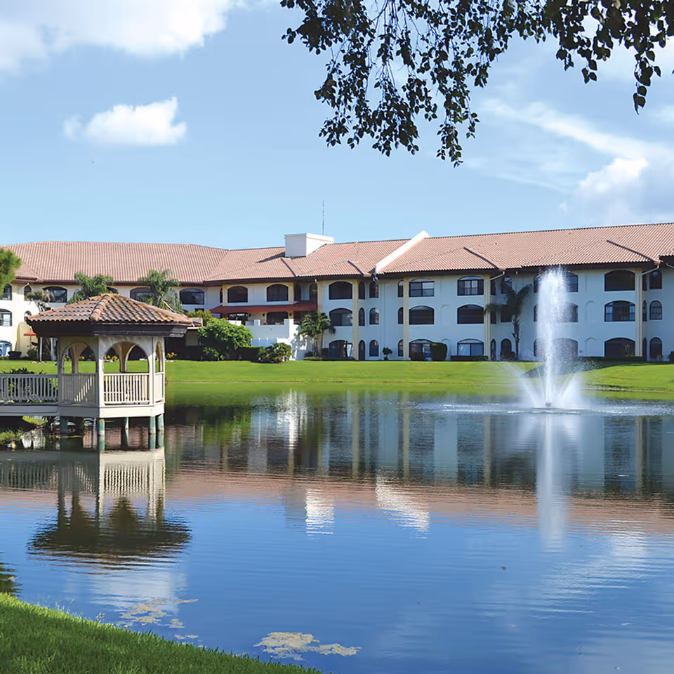 A large senior living facility building with a red-tiled roof and multiple windows, situated behind a pond with a water fountain and a small gazebo on the left side. The sky is clear with a few clouds and some tree branches hang from the top of the image.