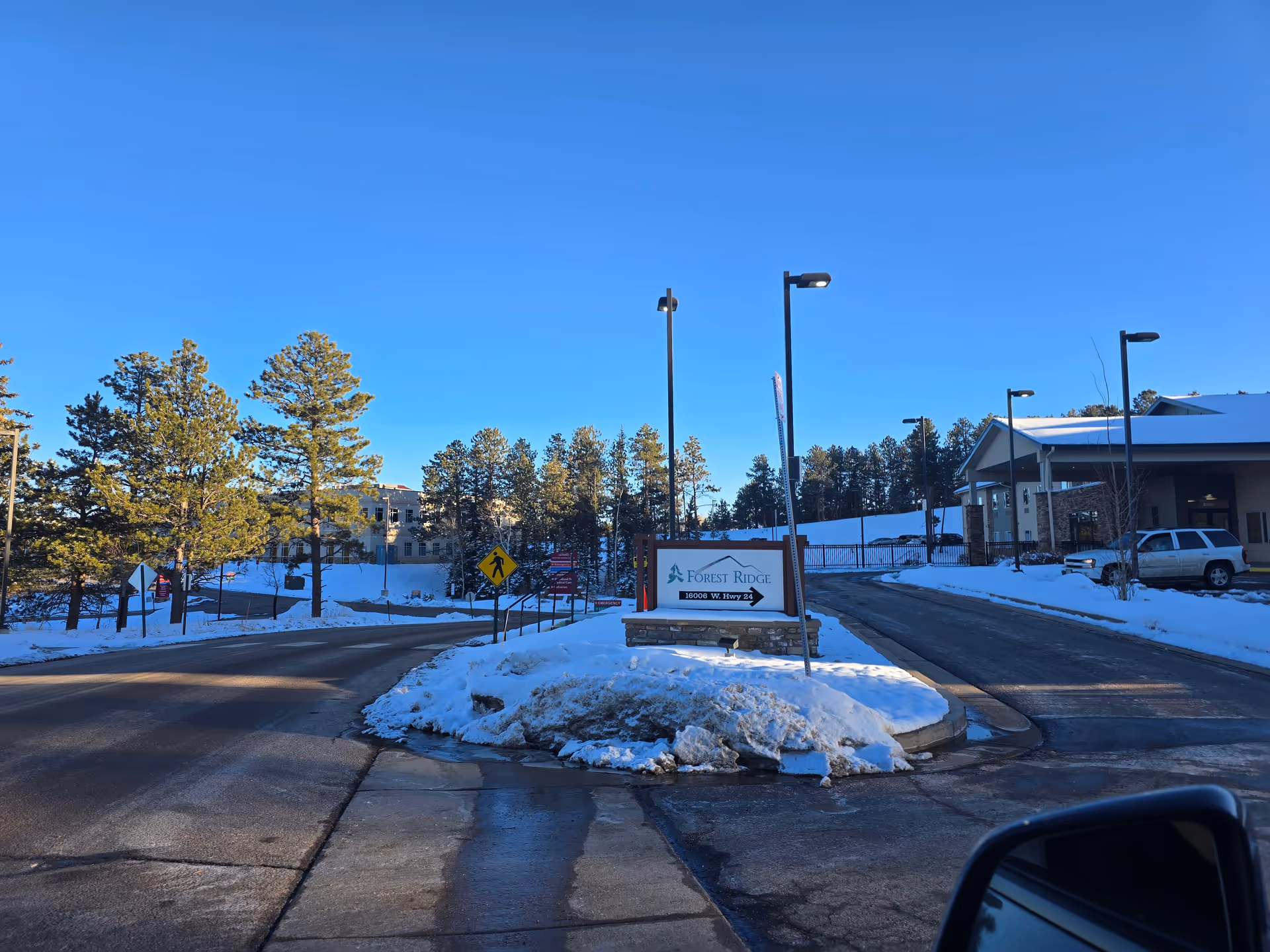Entrance road to Forest Ridge Senior Living facility with snow-covered ground, pine trees, street lamps, and a sign displaying the facility name and address under a clear blue sky.