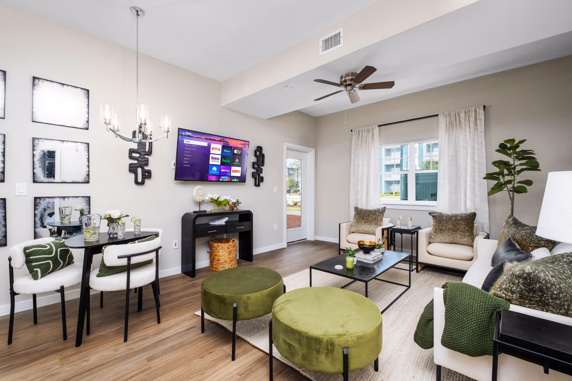 A modern living room with a small dining area. The living room features a white sofa with green and brown pillows, two green round ottomans, two beige armchairs, a black coffee table with decorative items, a ceiling fan, and a large window with white curtains. The dining area has a small black table with two white chairs, each with a green pillow. A flat-screen TV is mounted on the wall above a black console table with a basket underneath. The walls are light beige, and the floor is wood with a light-colored rug.