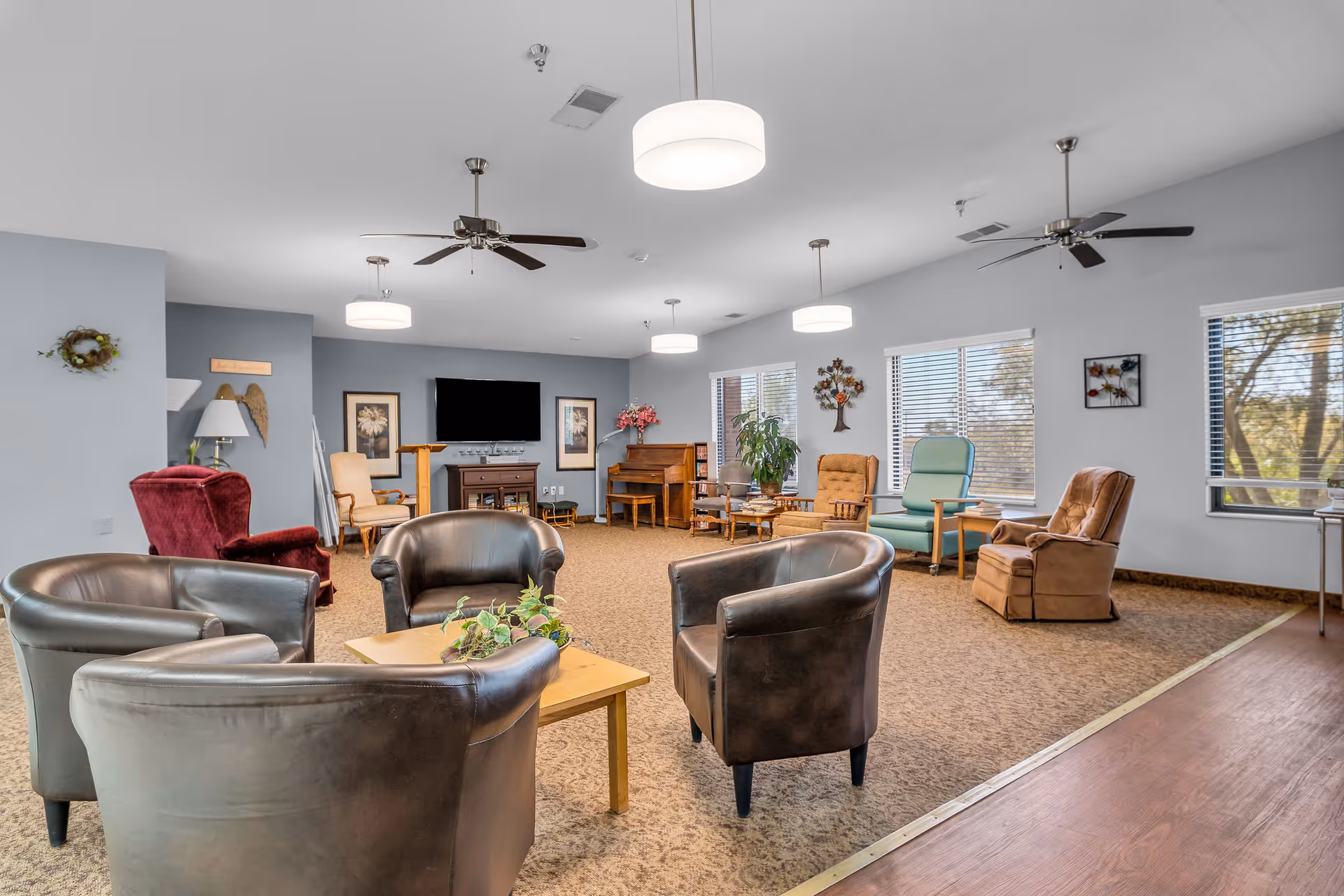Spacious senior living lounge with multiple armchairs arranged around a coffee table, a TV, piano, and large windows.