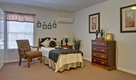 A cozy senior living bedroom with a single bed covered in a beige bedspread and a dark blue throw with white polka dots. There is a wooden armchair with patterned upholstery near a window with blinds and a valance. A wooden nightstand with a lamp and decorative items is beside the bed. On the opposite wall, there is a wooden chest of drawers with framed pictures, candles, and a plant. Two framed floral artworks hang above the chest of drawers. The room has beige carpet and white walls.