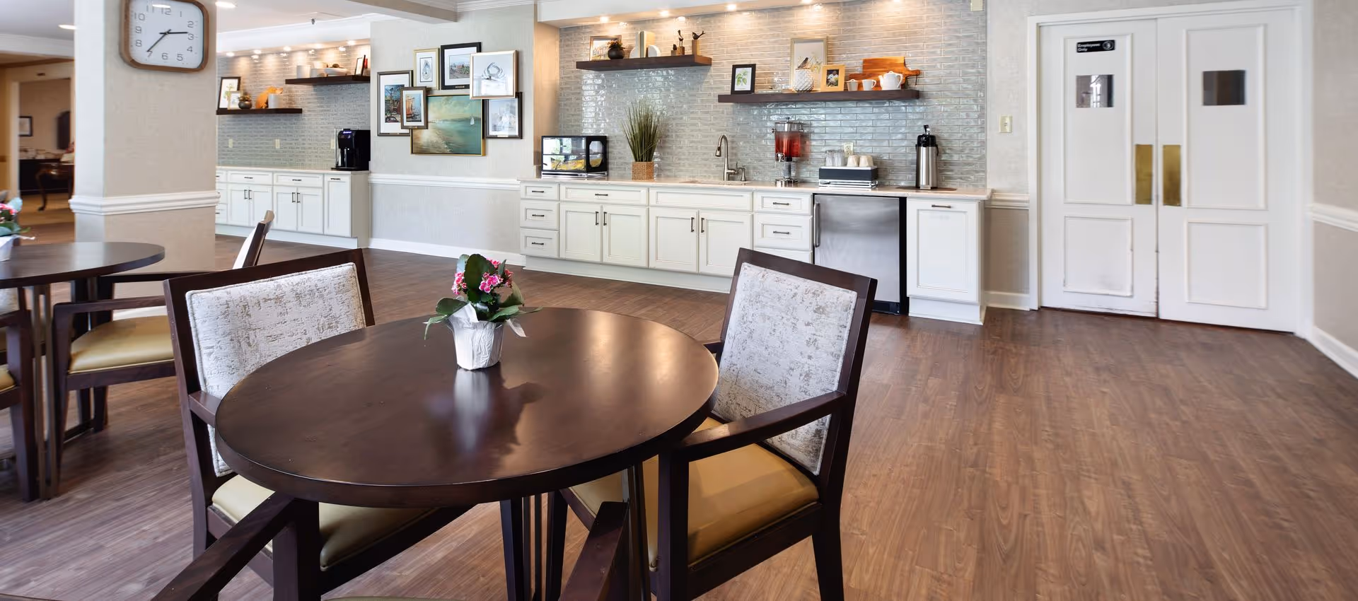 Bright senior living dining area with round wooden tables and chairs in front of a kitchenette with white cabinets and double doors.
