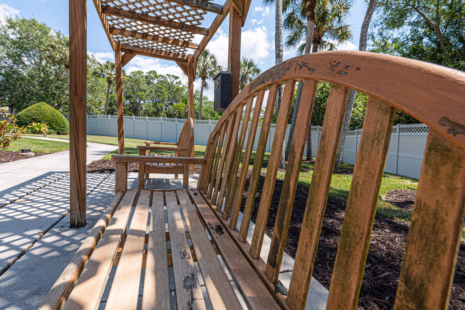 A wooden bench and chair set under a wooden pergola in an outdoor garden area with a concrete pathway, green grass, trees, and a white fence in the background under a blue sky with some clouds.