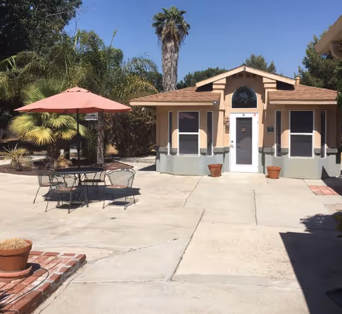 Outdoor patio area with a small table and four metal chairs under a red umbrella. The patio is paved with concrete and surrounded by potted plants and palm trees. In the background, there is a small beige building with a white door and windows, under a clear blue sky.