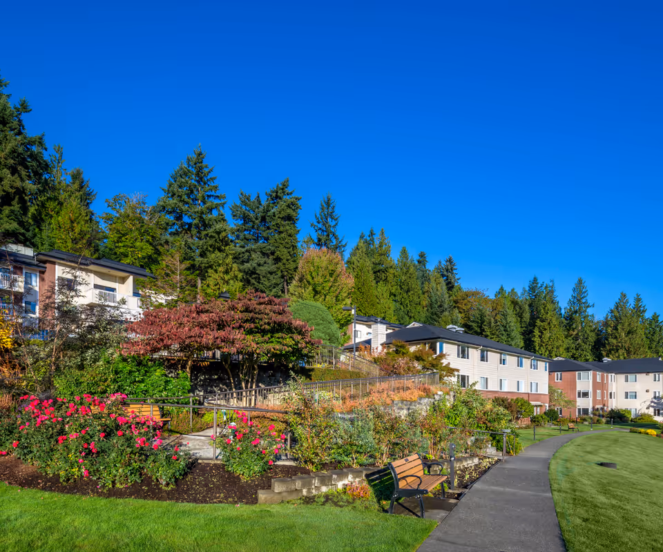 Outdoor view of Covenant Living at the Shores facility showing a landscaped garden with blooming flowers, a paved walking path, benches, and multi-story residential buildings surrounded by tall trees under a clear blue sky.