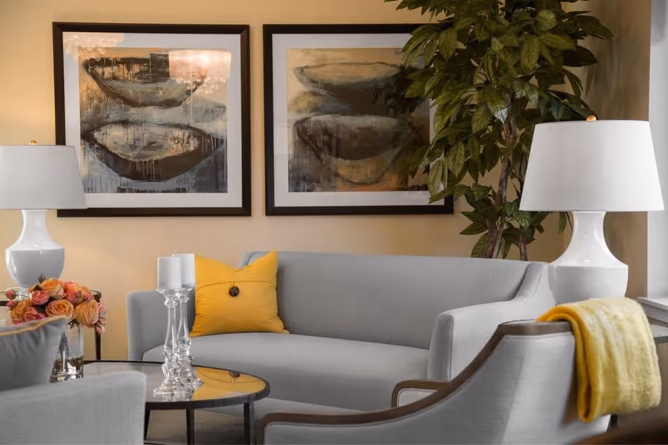 A cozy living room with a light gray sofa adorned with a yellow pillow, two white table lamps on either side, a glass coffee table with two white candles, a bouquet of flowers, two abstract paintings on the wall, and a large green potted plant in the corner.