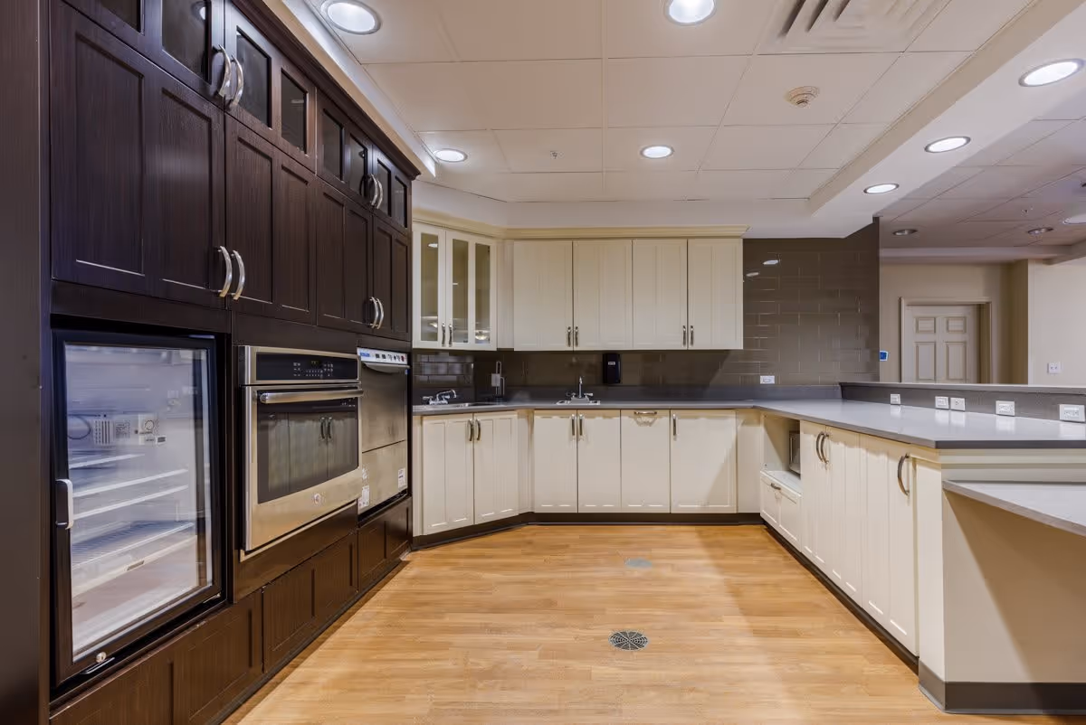 A spacious kitchen area with dark wood cabinets on the left side and light cream cabinets along the back and right walls. The kitchen features built-in appliances including an oven and a refrigerator. The floor is wooden, and the ceiling has recessed lighting. There is a countertop with multiple electrical outlets on the right side.