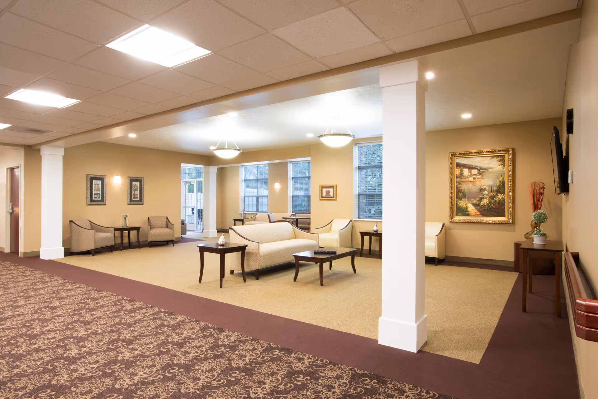 A spacious and well-lit common area in a senior living facility with beige walls and carpeted floors. The room features several cushioned chairs and sofas arranged around wooden coffee tables. Large windows allow natural light to enter, and the walls are decorated with framed artwork. Ceiling lights provide additional illumination, and there are white columns dividing the space.