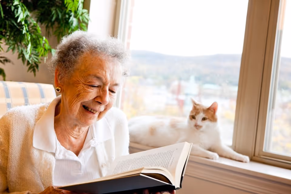 An elderly woman with short curly gray hair and gold earrings is smiling while reading a book. She is sitting on a couch with a plaid pattern near a large window. A white and orange cat is lying on the windowsill behind her, with a blurred outdoor view visible through the window.