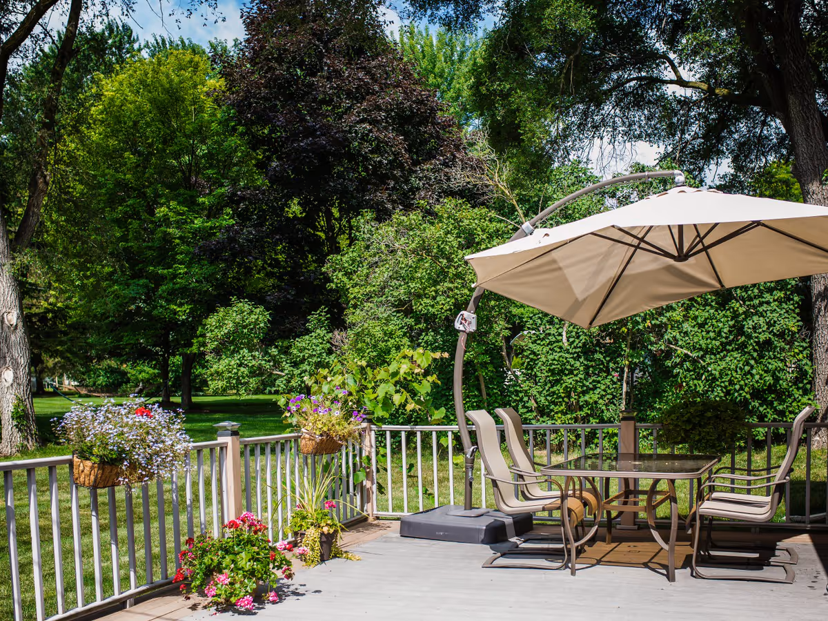 Outdoor patio area with a glass-top table and four chairs under a large beige umbrella. The patio is surrounded by a white railing with hanging flower baskets and potted plants. In the background, there are lush green trees and a well-maintained lawn under a clear blue sky.