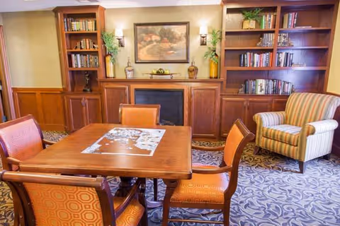Cozy senior living common room with a wooden table and chairs, built-in bookshelves, a fireplace, and an upholstered armchair.